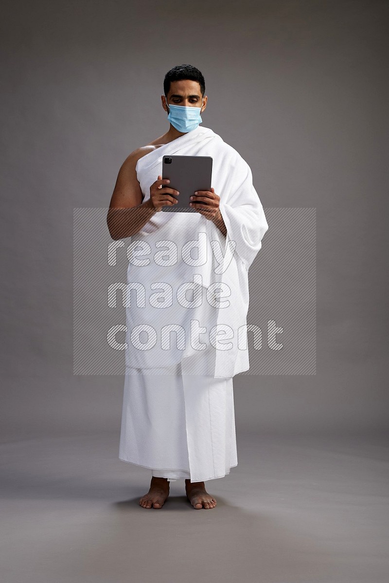 A man wearing Ehram with face mask Standing holding a tablet on gray background