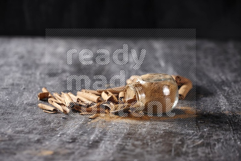 Herbal glass jar full cinnamon powder flipped and a metal spoon full of powder surrounded by cinnamon sticks on textured black background in different angles