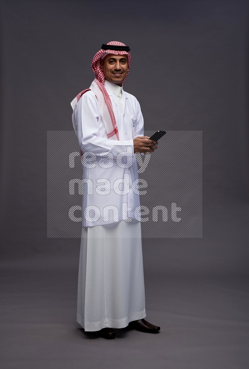 Saudi man wearing thob with lab coat and shomag with pocket employee badge standing texting on phone on gray background