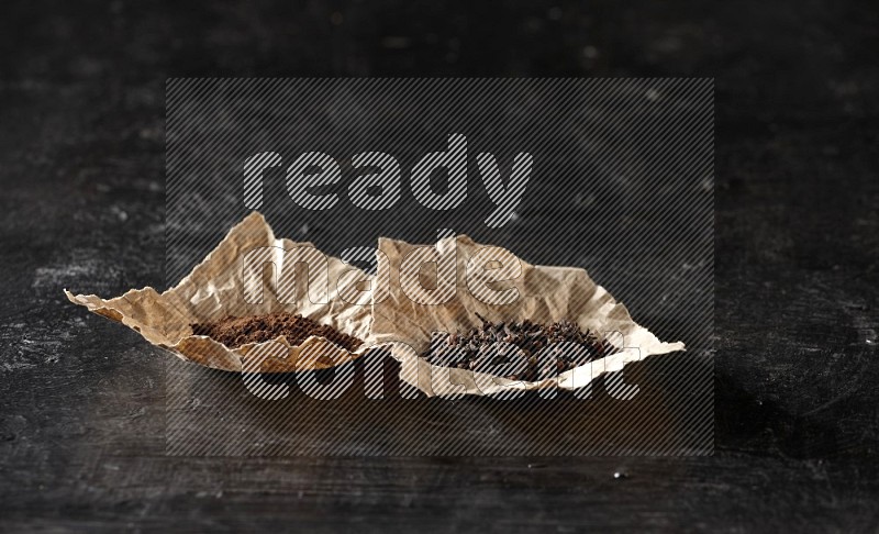 Cloves powder and cloves in 2 crumpled pieces of paper on a textured black flooring