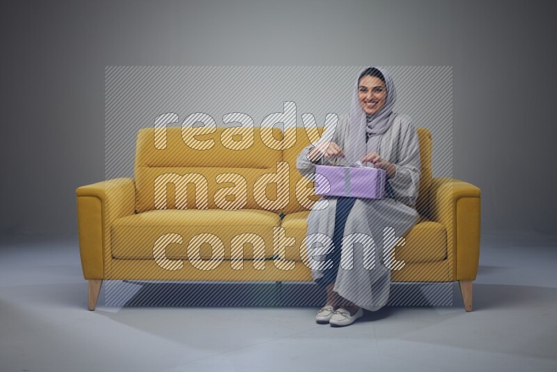 A Saudi woman wearing a light gray Abaya and head scarf sitting on a yellow sofa holding a gift box beside multi color gift boxes eye level on a grey background
