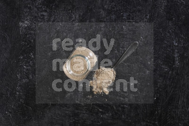 A glass spice jar full of garlic powder with a filled metal spoon on a textured black flooring