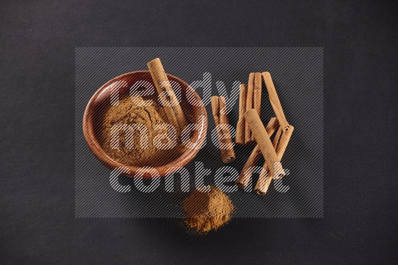 Cinnamon powder in a wooden bowl with a cinnamon sticks on black background