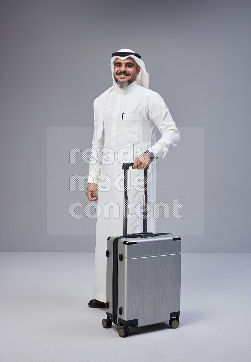 Saudi man wearing Thob and white Shomag standing holding Travel bag on Gray background