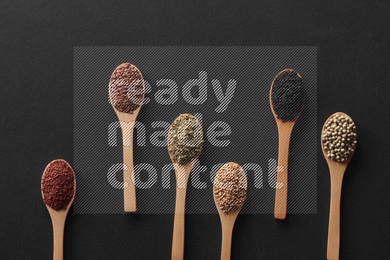 wooden spoons filled with white peppers, mustard seeds, black seeds, garden cress, cumin and flax on black flooring