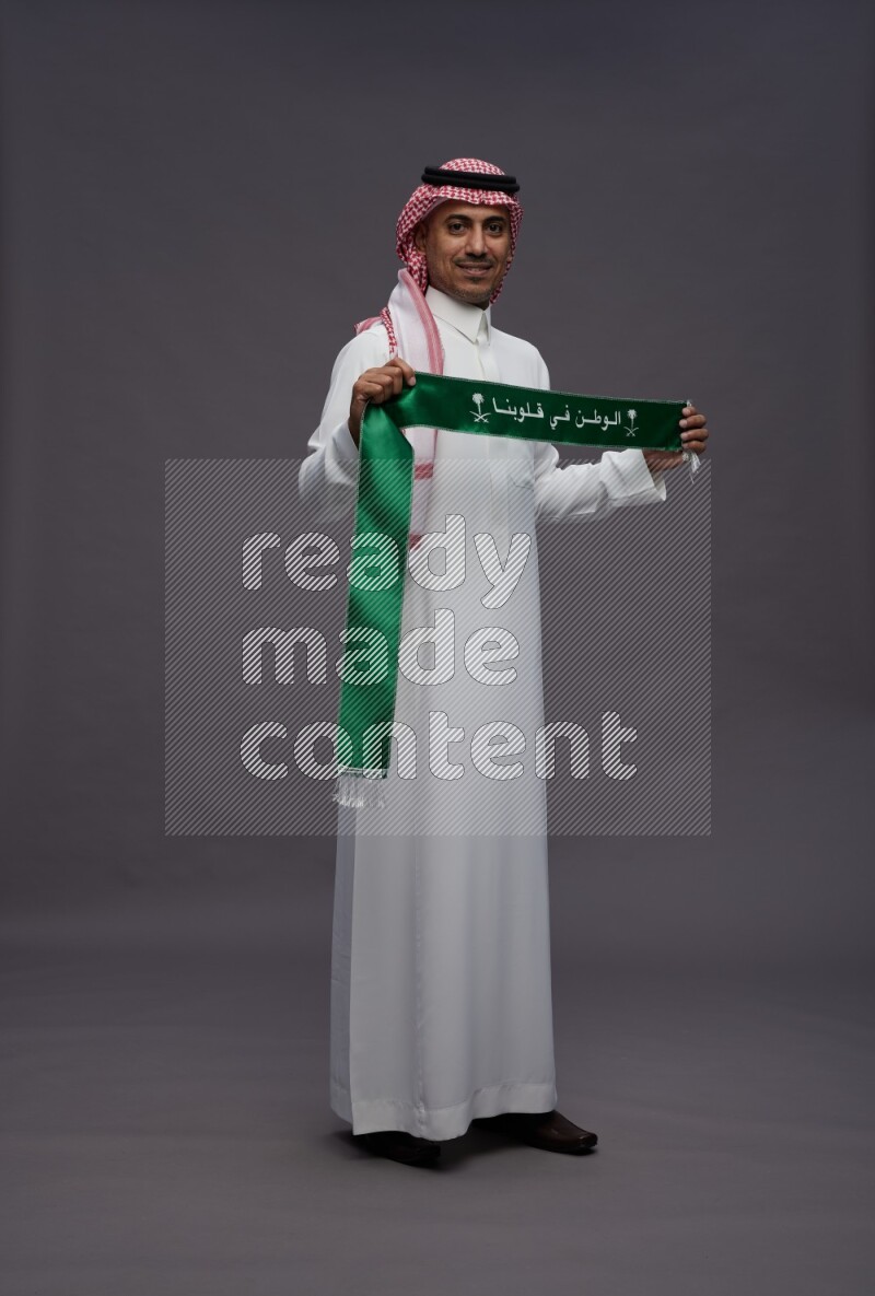 Saudi man wearing thob and shomag standing holding Saudi flag on gray background