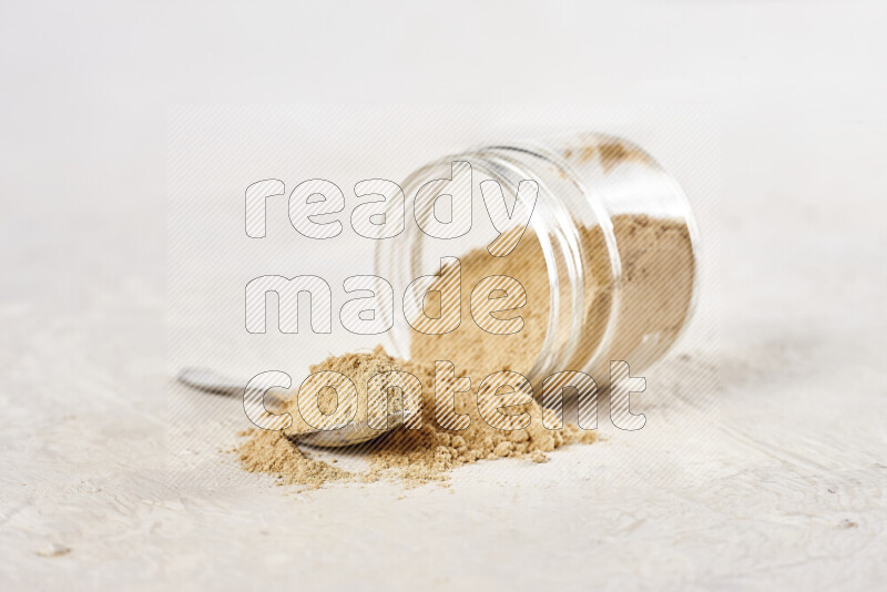 A glass jar full of ground ginger powder flipped with some spilling powder on white background