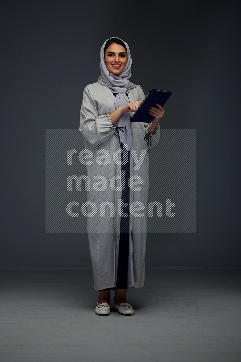 A Saudi woman wearing a light gray Abaya and head scarf standing holding a phone and pointing with the other hand eye level on a grey background