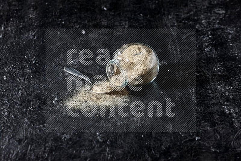 A flipped herbal glass jar and a metal spoon full of white pepper powder with spilled powder on textured black flooring