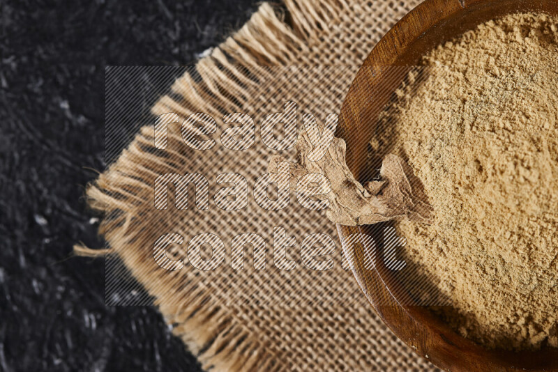 A wooden bowl full of ground ginger powder with a wooden spoon on it all on a burlap fabric on black background