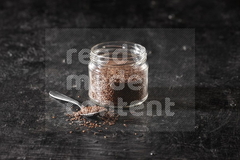 A glass jar full of flax with a metal spoon full of the seeds on a textured black flooring in different angles