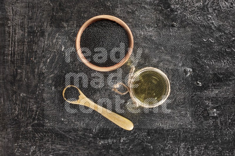 A wooden bowl and spoon full of black seeds and a glass jar of black seeds oil on a textured black flooring