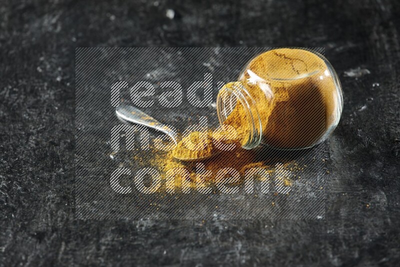 A flipped glass spice jar and a metal spoon full of turmeric powder and powder spilled out of it on textured black flooring