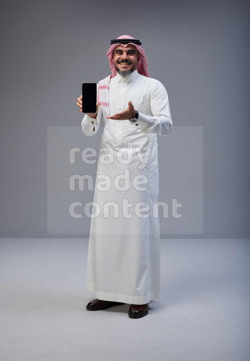 Saudi man Wearing Thob and red Shomag standing showing phone to camera on Gray background