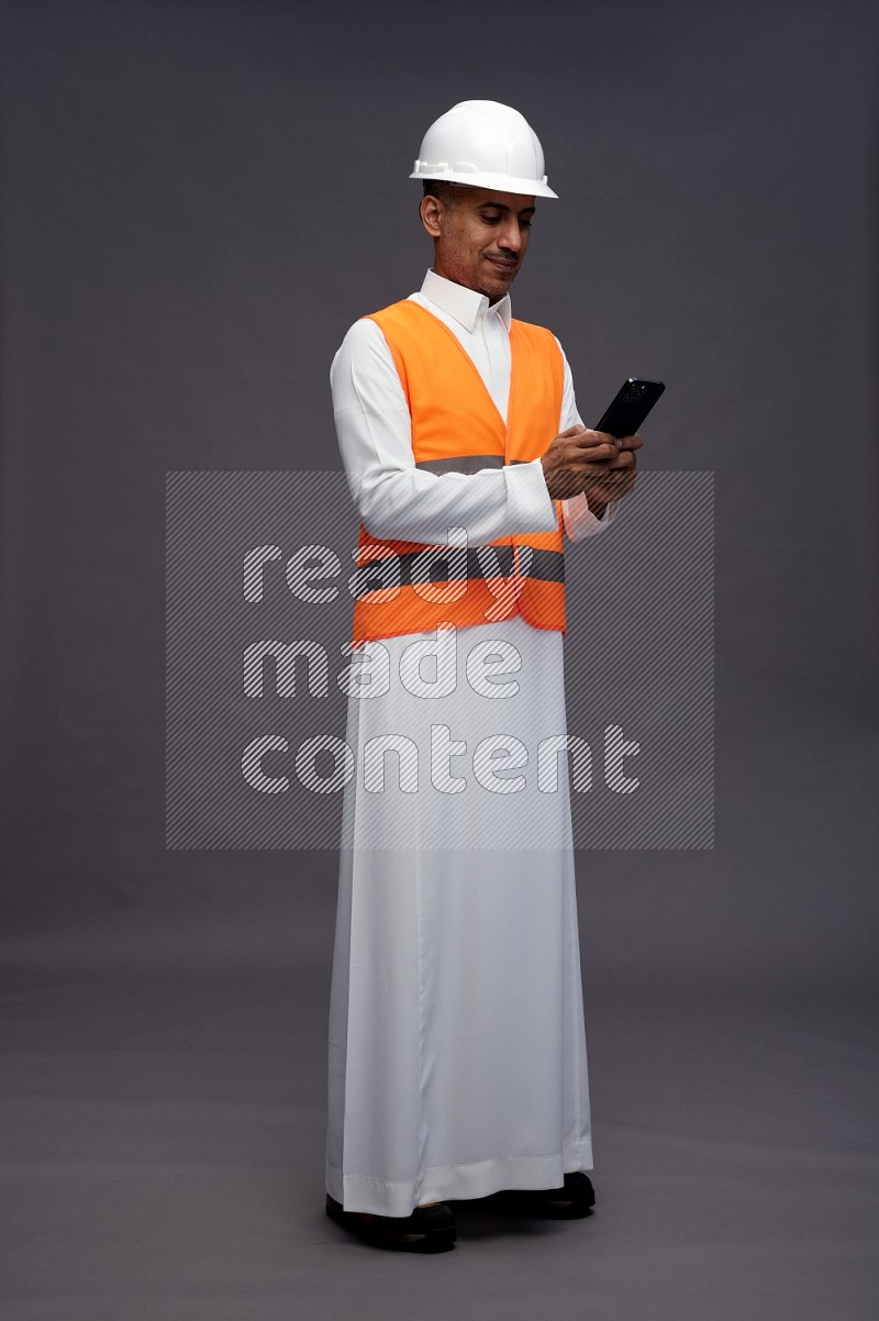 Saudi man wearing thob with engineer vest standing texting on phone on gray background