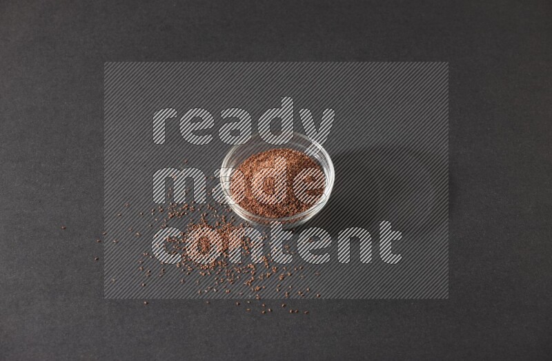 A glass bowl full of garden cress seeds surrounded by seeds on a black flooring