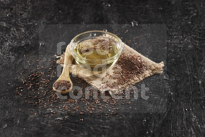 A glass bowl full of flaxseeds oil and wooden spoon full of flaxseeds with seeds spread on burlap fabric on a textured black flooring