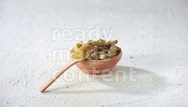A wooden spoon full of turmeric powder above a wooden bowl full of dried turmeric whole fingers on a textured white flooring