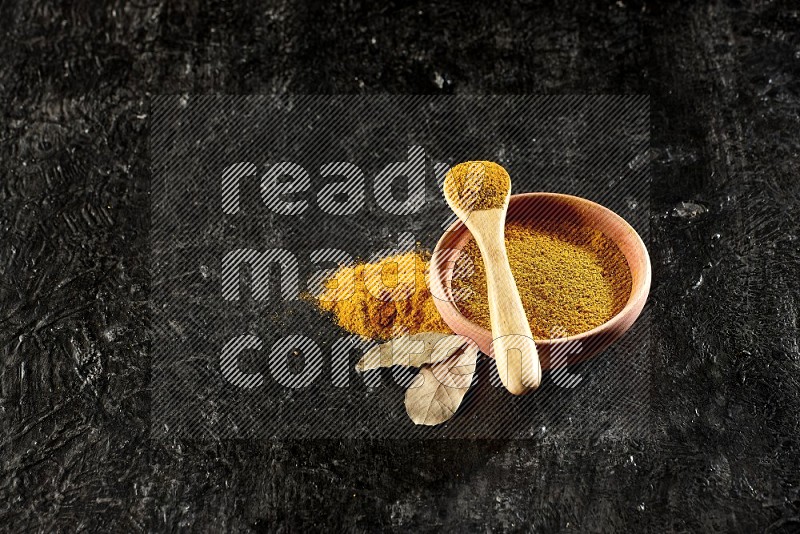 A wooden bowl and a wooden spoon full of turmeric powder on textured black flooring