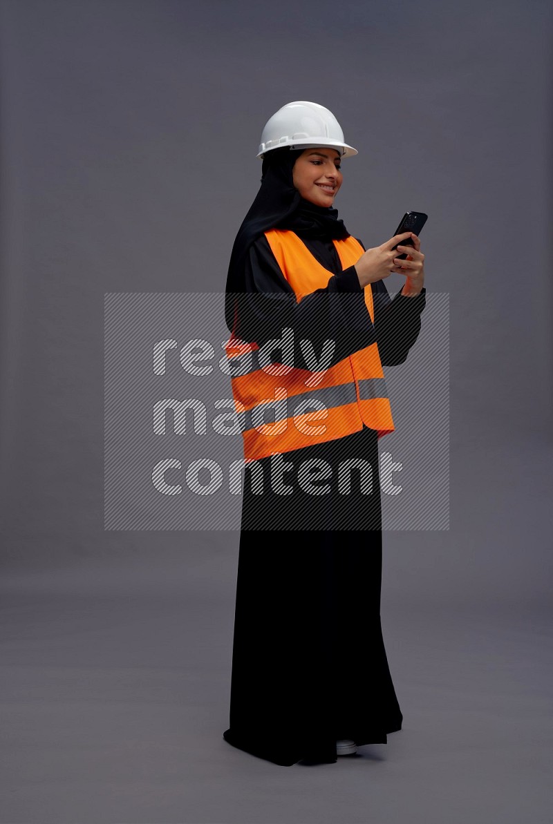 Saudi woman wearing Abaya with engineer vest standing texting on phone on gray background