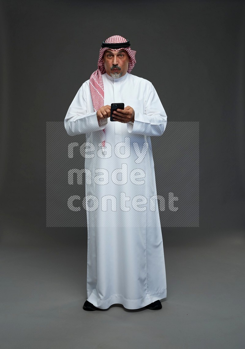 Saudi man with shomag Standing texting on phone on gray background