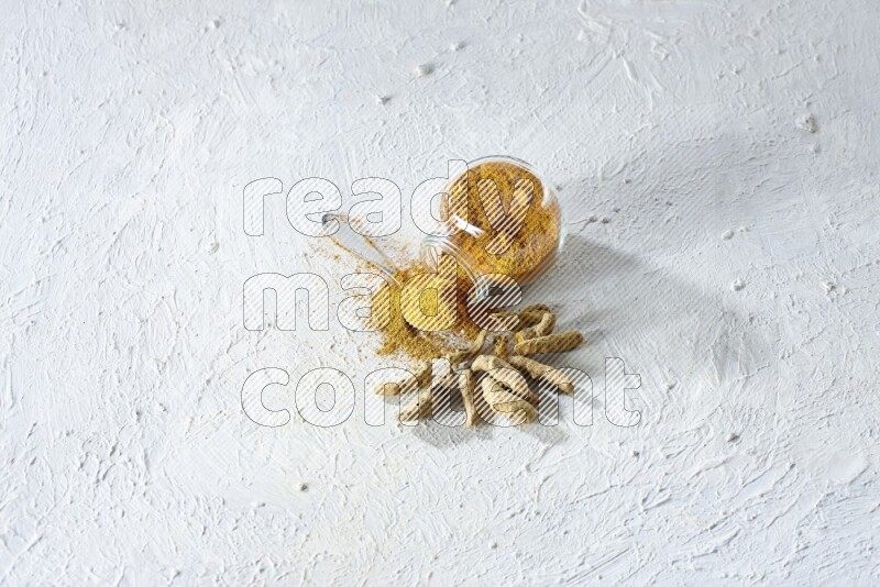 A flipped glass spice jar and metal spoon full of turmeric powder and powder spilled out of it with dried whole fingers on textured white flooring