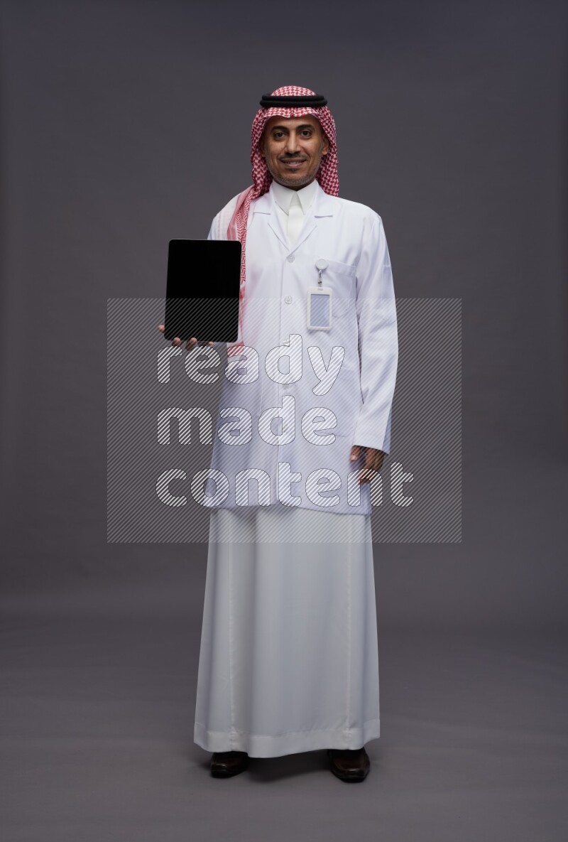 Saudi man wearing thob with lab coat and shomag with pocket employee badge standing showing tablet to camera on gray background