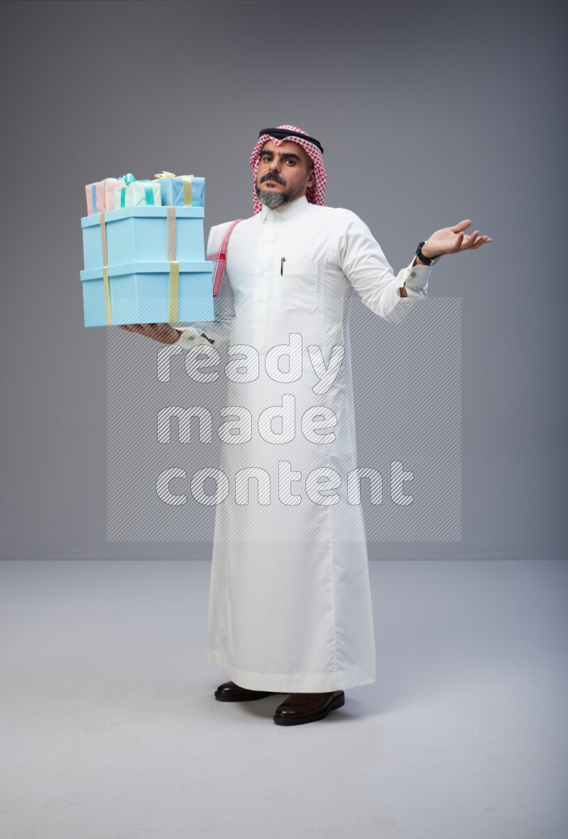 Saudi man Wearing Thob and red Shomag standing holding gift box on Gray background