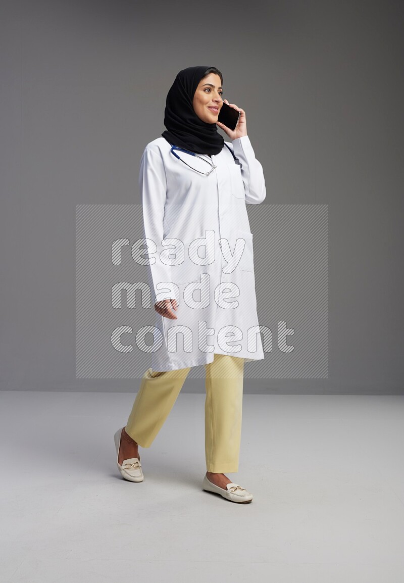 Saudi woman wearing lab coat with stethoscope standing talking on phone on Gray background