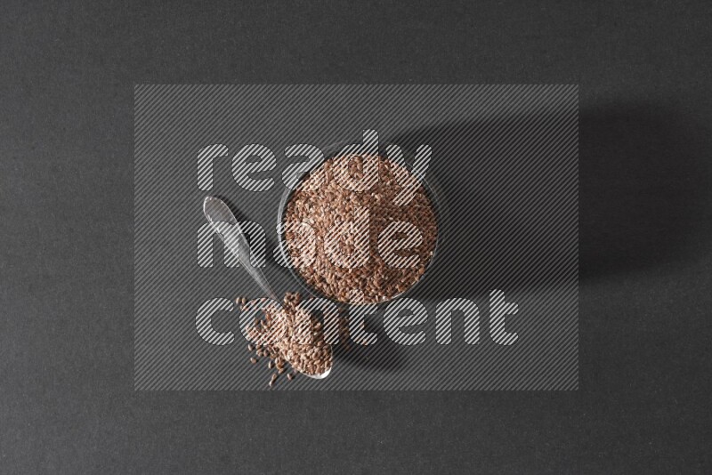 A black pottery bowl full of flaxseeds and a metal spoon full of the seeds on a black flooring