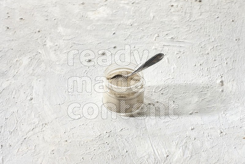 وعاء زجاجي وملعقة معدنية ممتلئان ببودرة الفلفل الأبيض على أرضية بيضاء