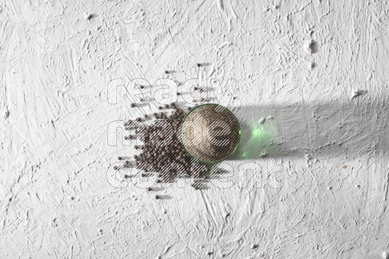 فلفل أسود مطحون في كوب زجاجي مع بعض حبوب الفلفل علي خلفية بيضاء