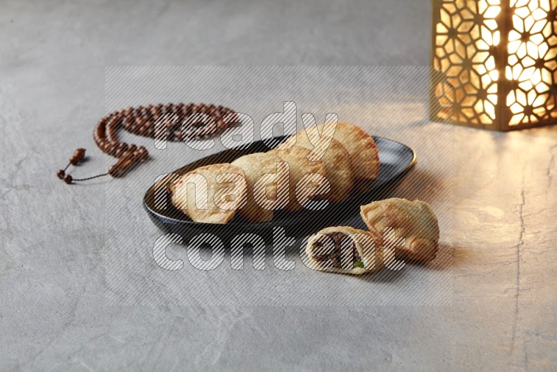 Five fried sambosas in an oval shaped black plate, beside a cut meat sambosa, a brown misbaha and a golden lantern on a gray background