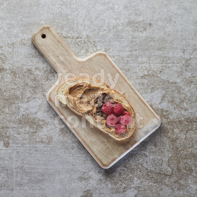 open faced peanut butter sandwich with raspberries and chocolate granula on a grey textured background