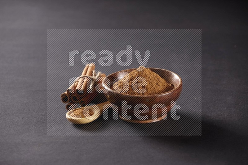 Cinnamon sticks stacked and bounded beside a wooden bowl full of cinnamon powder and a wooden spoon full of powder on black background