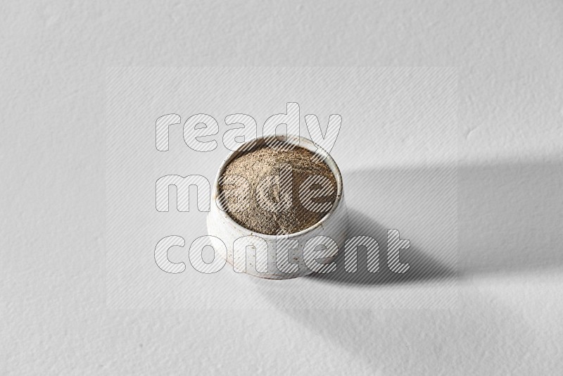 A white ceramic bowl full of black pepper powder and black pepper beads spread on white flooring