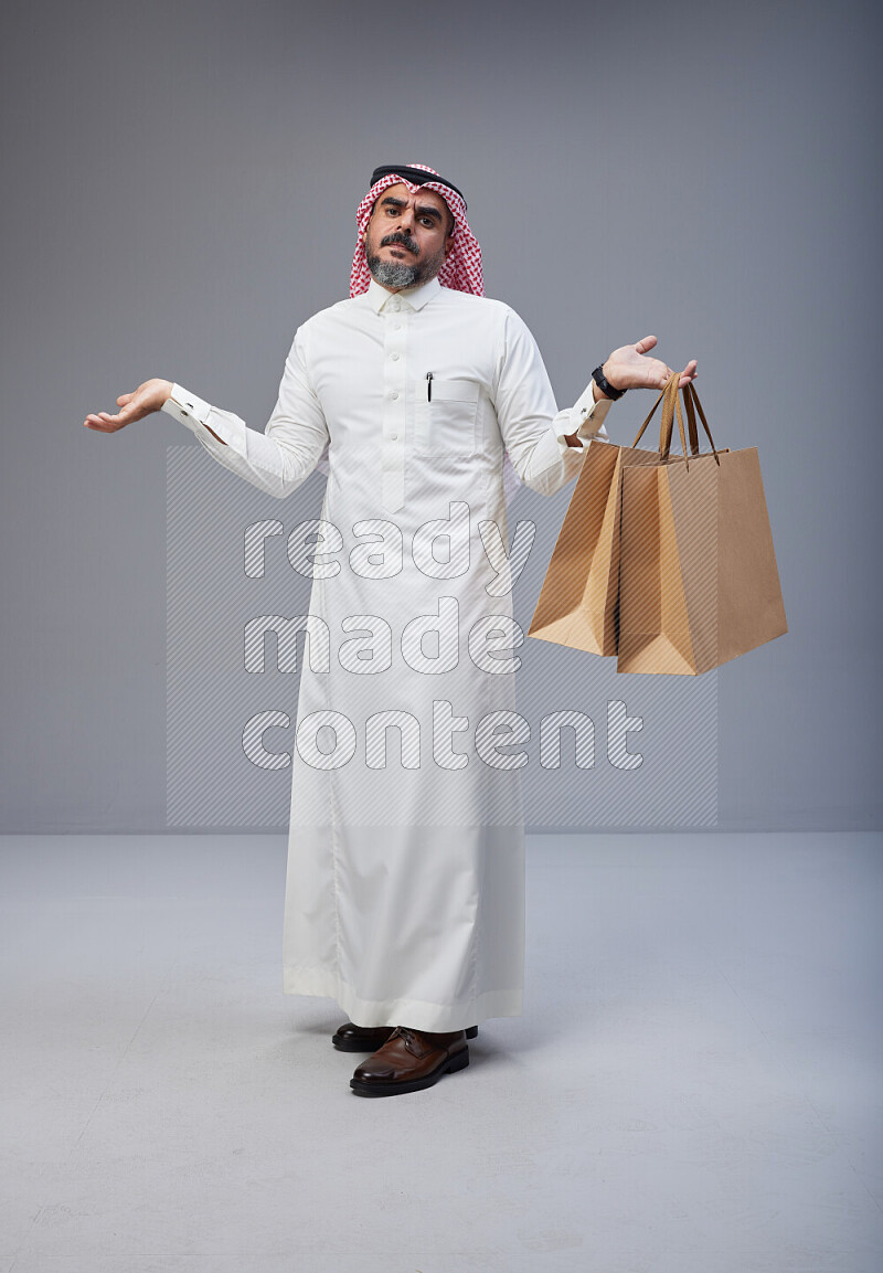 Saudi man Wearing Thob and red Shomag standing holding shopping bag on Gray background