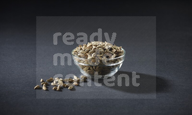 A glass bowl full of cardamom and more seeds spreaded beneath the bowl on black flooring