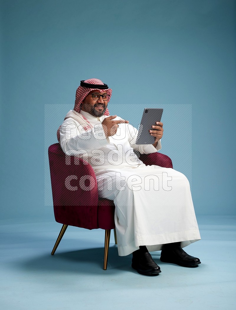 Saudi Man with shimag sitting on chair working on tablet on blue background