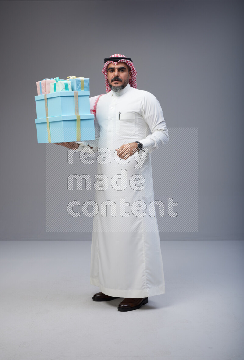 Saudi man Wearing Thob and red Shomag standing holding gift box on Gray background