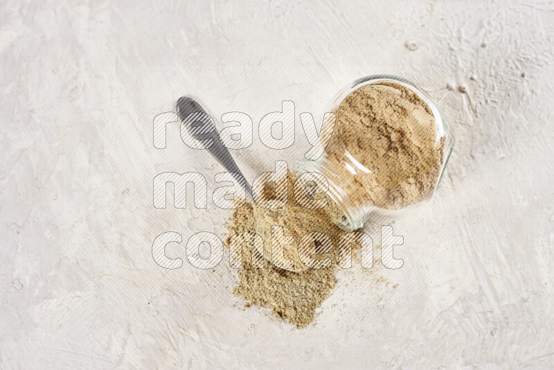 A glass jar full of ground ginger powder flipped with some spilling powder on white background