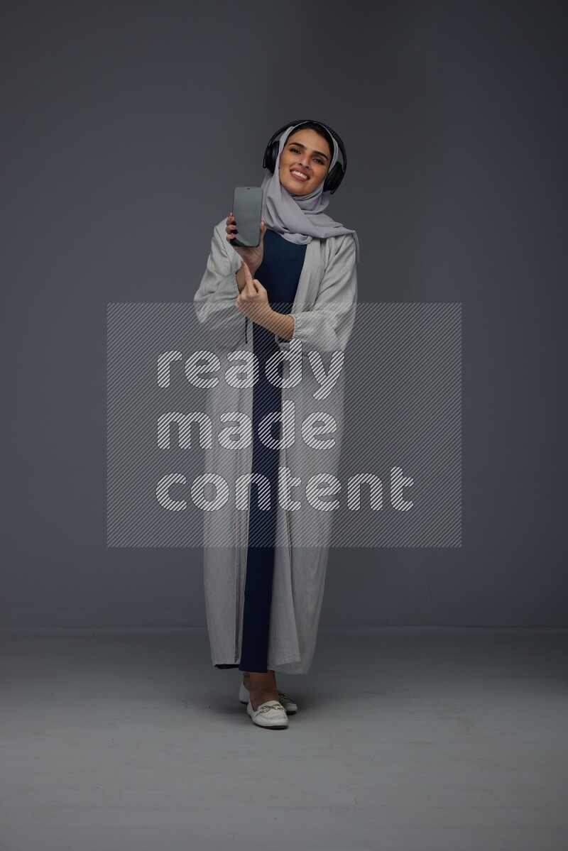 A Saudi woman wearing a light gray Abaya and head scarf setting on a dark grey chair and watching on the tablet eye level on a grey background