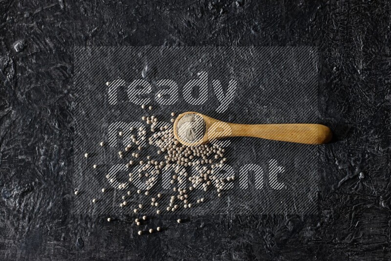 A wooden spoon full of white pepper powder with white pepper beads on textured black flooring