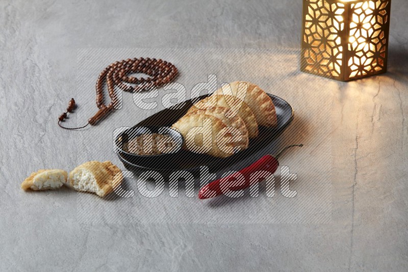 Four fried sambosas in an oval shaped black plate, beside a cut cheese sambosa, a brown misbaha and a golden lantern on a gray background