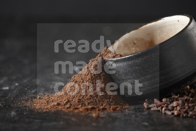 A black pottery bowl full of cloves powder spilled out of it with whole grains on a textured black flooring