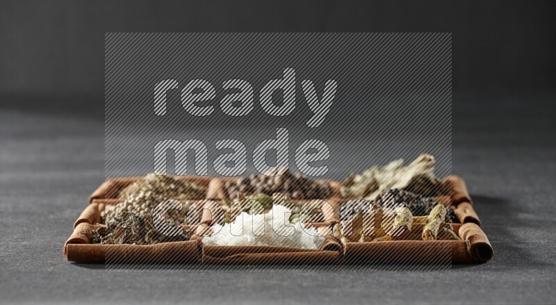 9 squares of cinnamon sticks full of cardamom in the middle surrounded by turmeric, black peppers, bay laurel, allspice, cumin, white peppers, basil and salt on black flooring