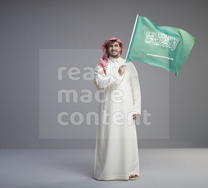 A Saudi man standing wearing thob and red shomag raising big Saudi flag on gray background