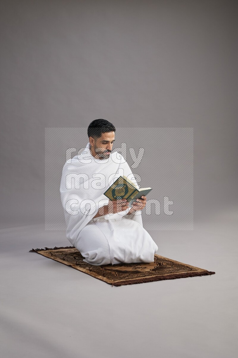 A man wearing Ehram sitting on floor reading quran on gray background