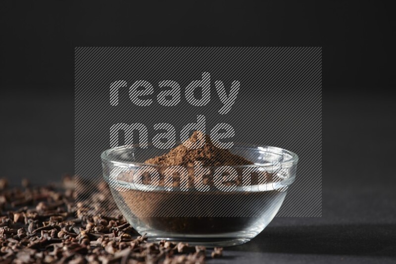 وعاء زجاجي ممتلئ ببودرة القرنفل مع حبوب القرنفل المتناثرة على أرضية سوداء