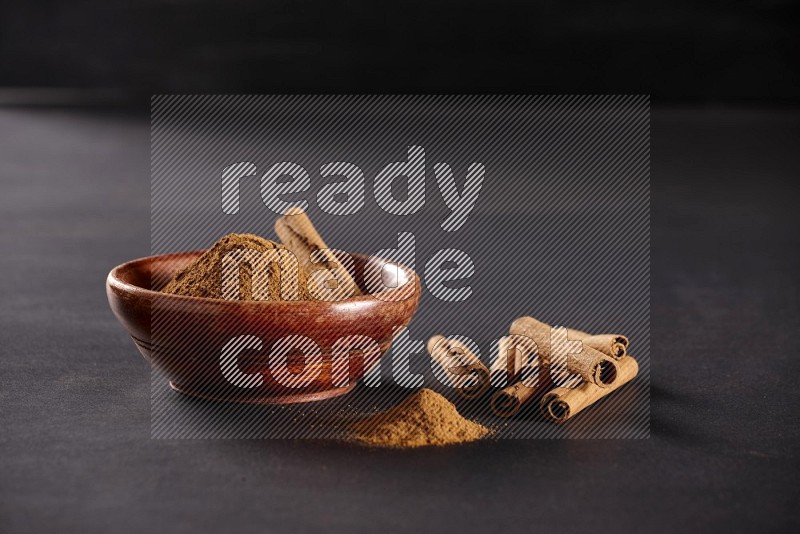 Cinnamon powder in a wooden bowl with a cinnamon sticks on black background